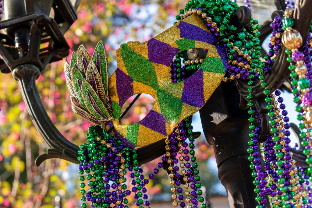 Mardi Gras mask with vibrant purple, green, and gold beads draped over a black street lamp in New Orleans, symbolizing the city’s festive spirit.