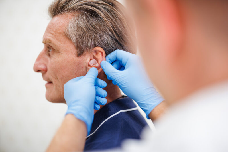 An audiologist in blue gloves fits a clear custom earmold into a man's ear.